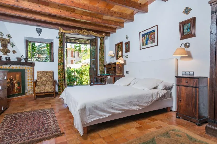 A bedroom with a bed, exposed wooden ceiling beams, and a window overlooking greenery.