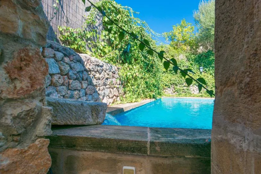 A bright swimming pool is seen beyond a stone opening, surrounded by greenery.