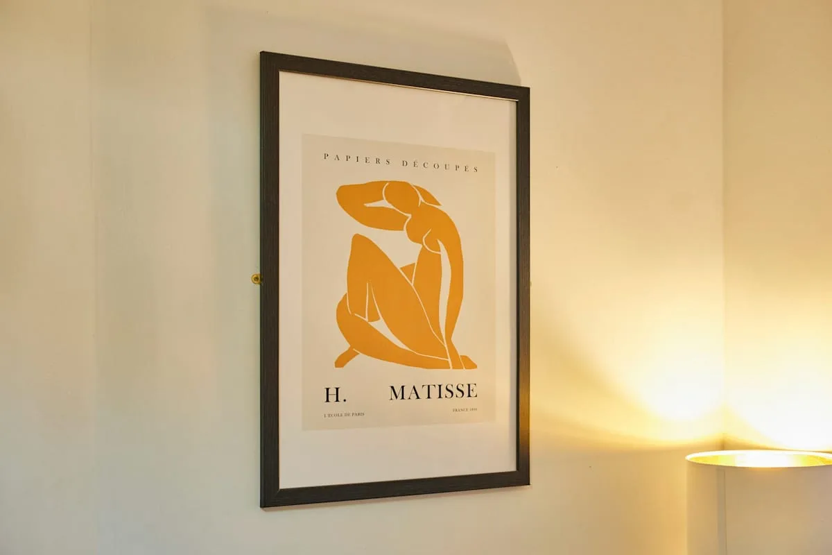 A framed art print hangs on a light-colored wall beside a glowing lamp.