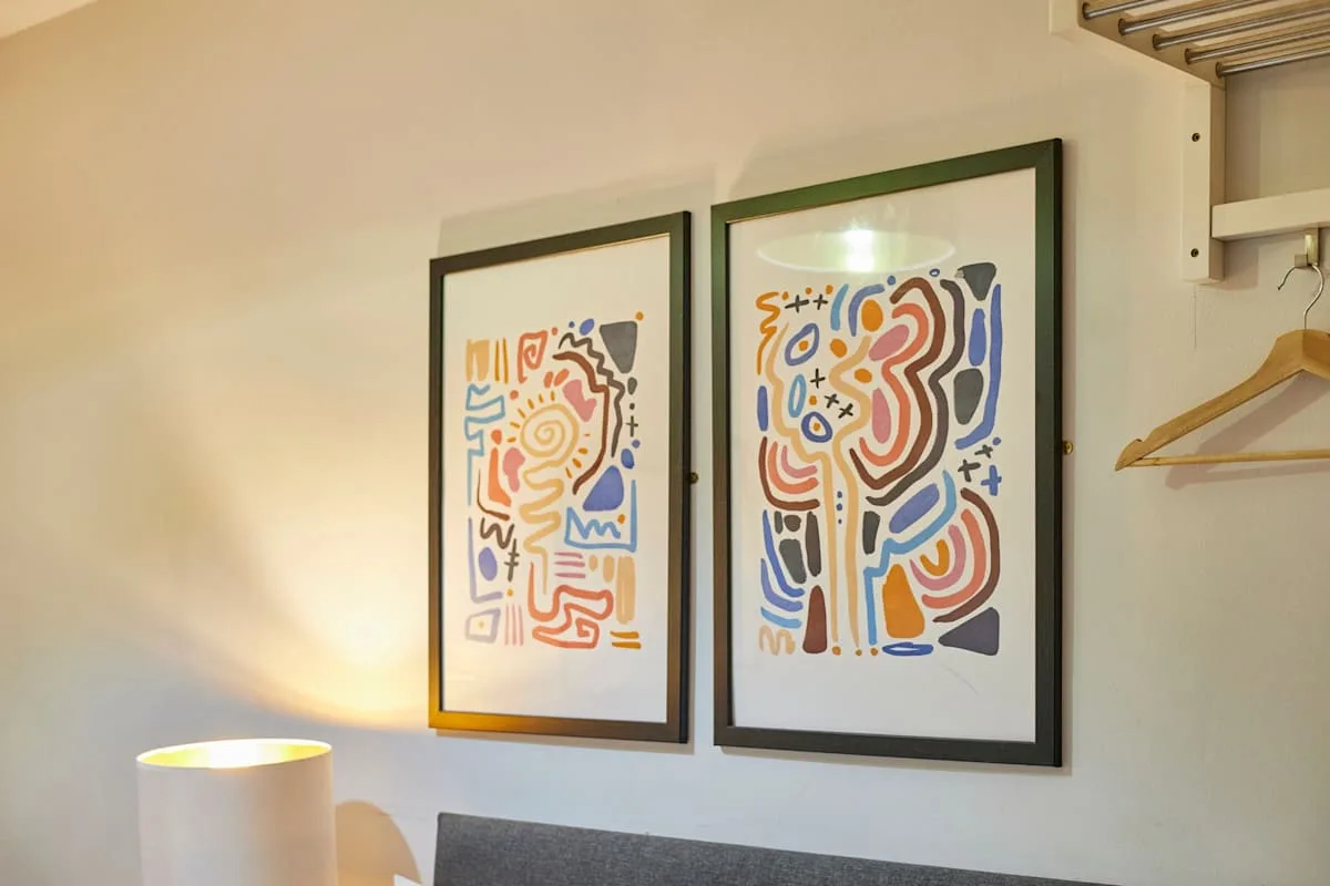 A bedroom wall displays two colorful abstract prints above a bedside lamp.