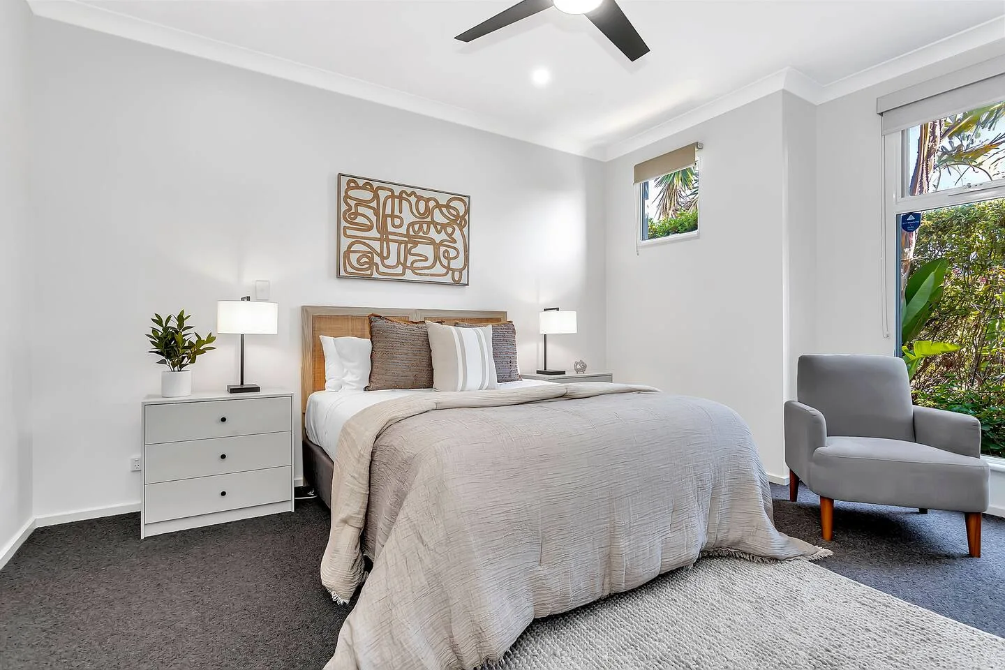 Un dormitorio con una cama bien tendida y una silla gris junto a la ventana.