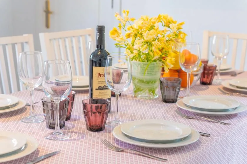 A dining table is set with plates, glasses, wine, and a vase of yellow flowers.