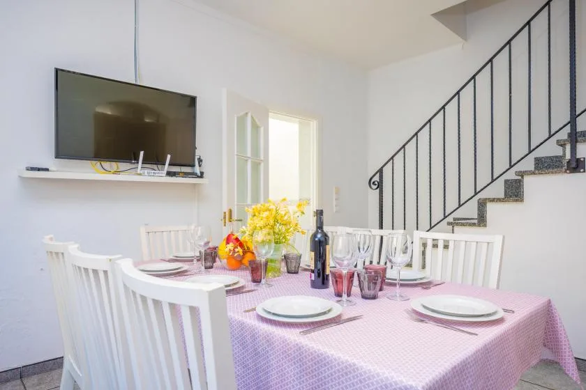 A dining table with a pink tablecloth is set beside a staircase and wall-mounted television.