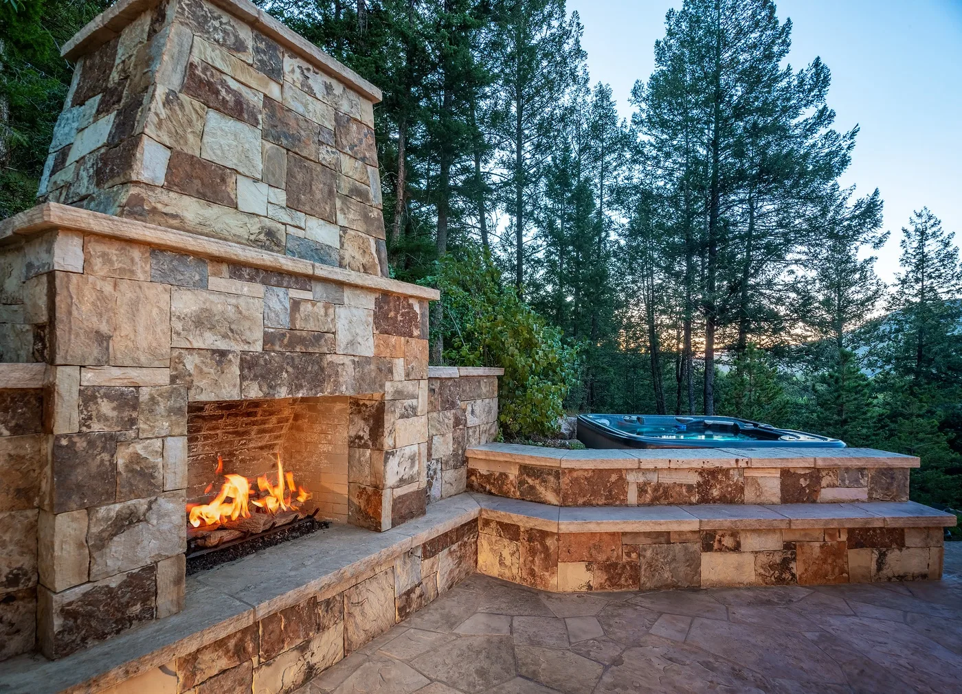 Un patio exterior cuenta con una chimenea de piedra encendida junto a una piscina.