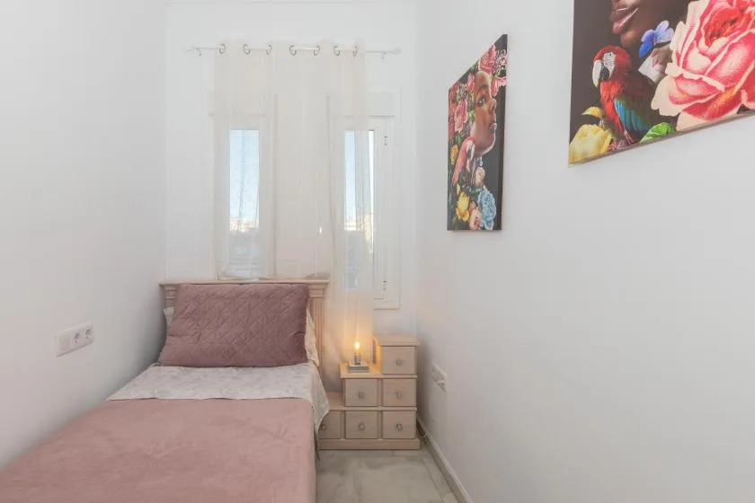 A narrow bedroom with a single bed, sheer curtains, and colorful wall art.