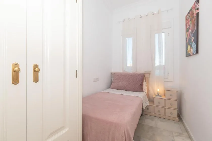 A bedroom with a bed, pale curtains, and a small bedside cabinet.