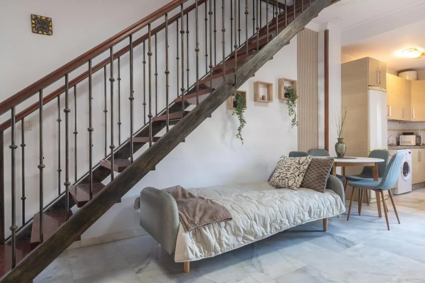 A living area with a daybed beneath a staircase, beside a dining table and kitchen.