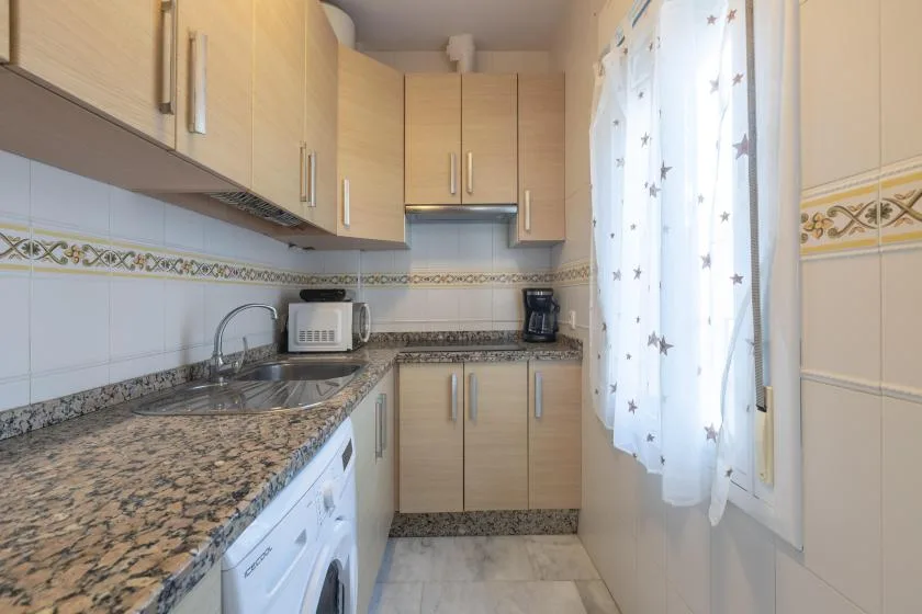 A kitchen with light wood cabinets, granite counters, and a window with sheer curtains.
