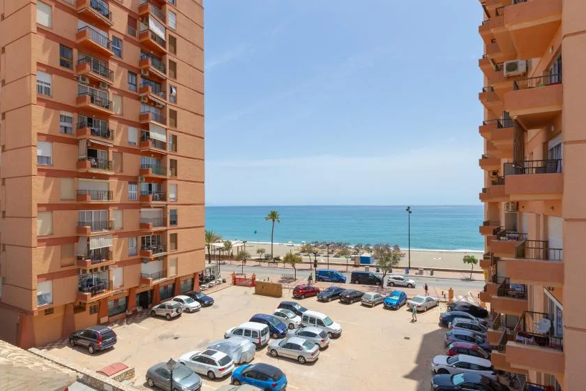 A view between apartment buildings overlooks a parking area and the sea.