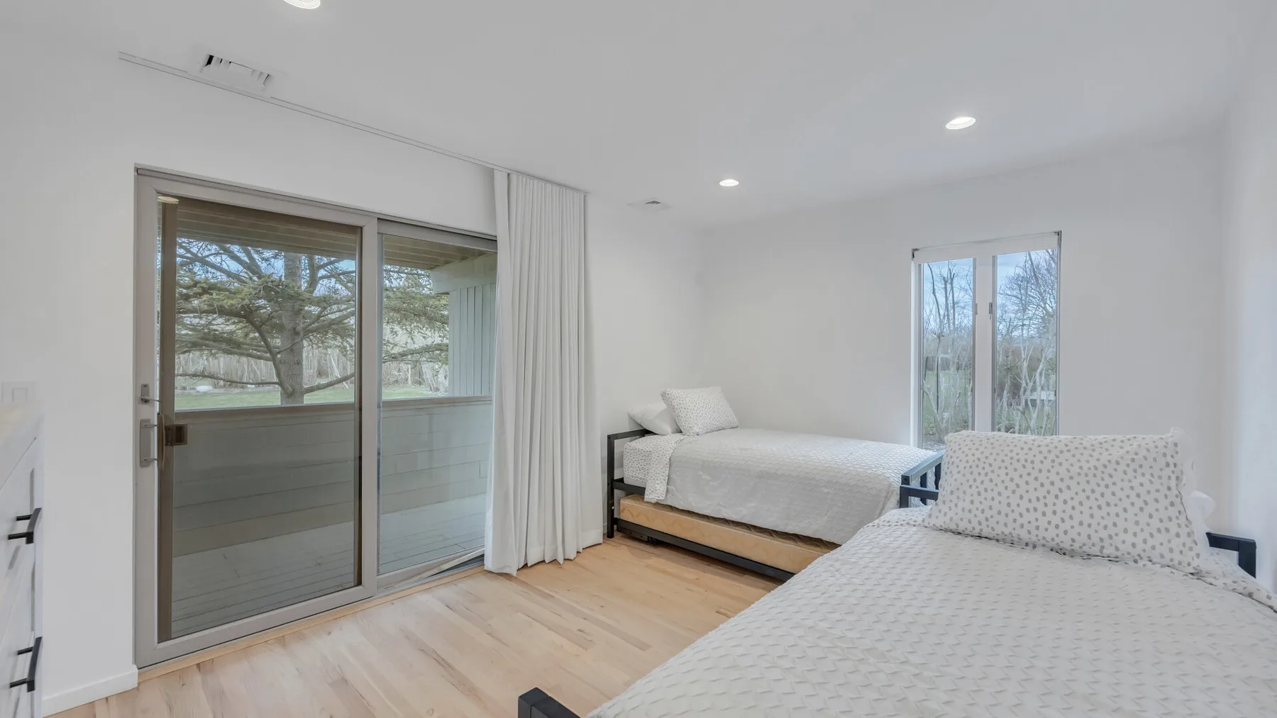 A bright bedroom has two beds and sliding glass doors opening onto a view of trees.