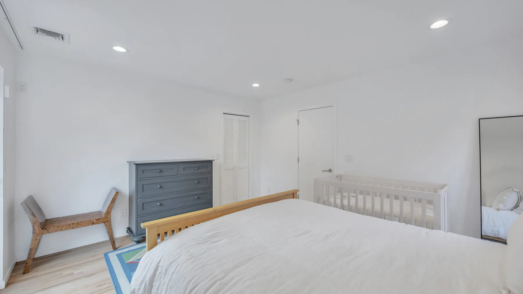 A bright bedroom with a large bed, a wooden crib, and a dresser.