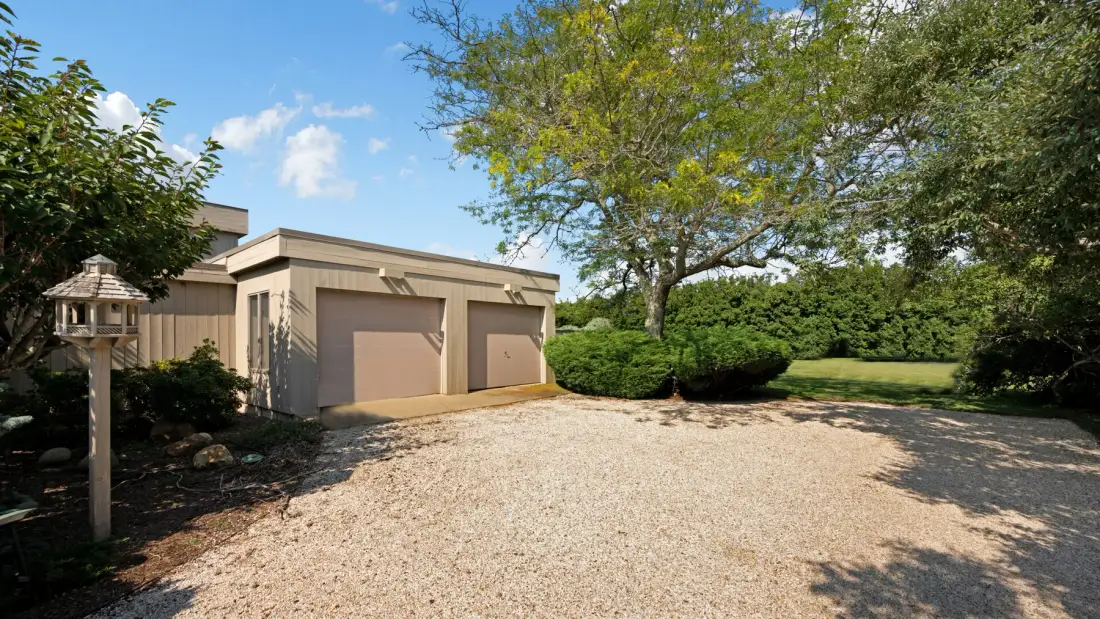A gravel driveway leads to a beige building with two garage doors, surrounded by trees and hedges.