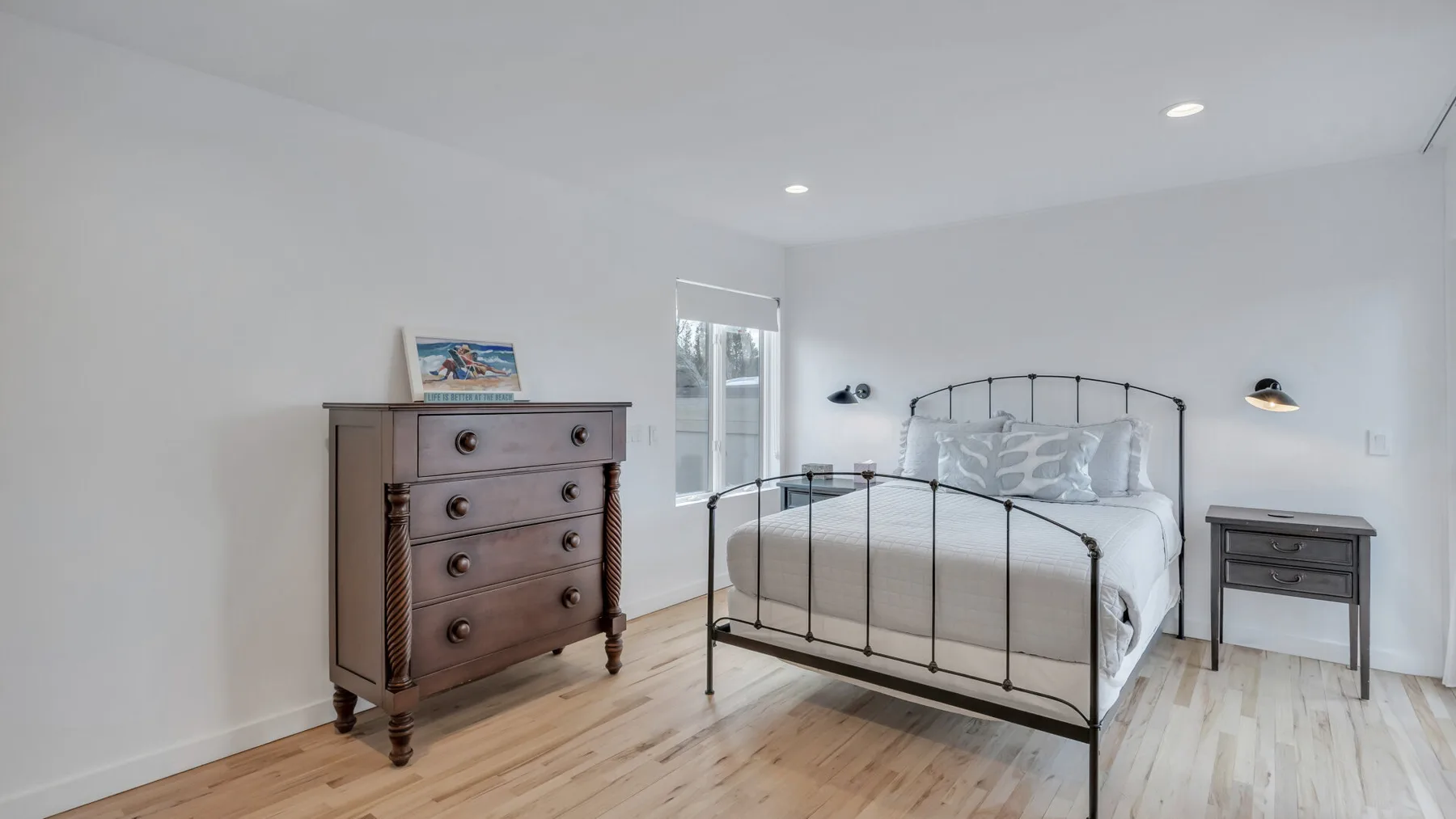 A bright bedroom with a metal bed, wooden dresser, and light wood flooring.