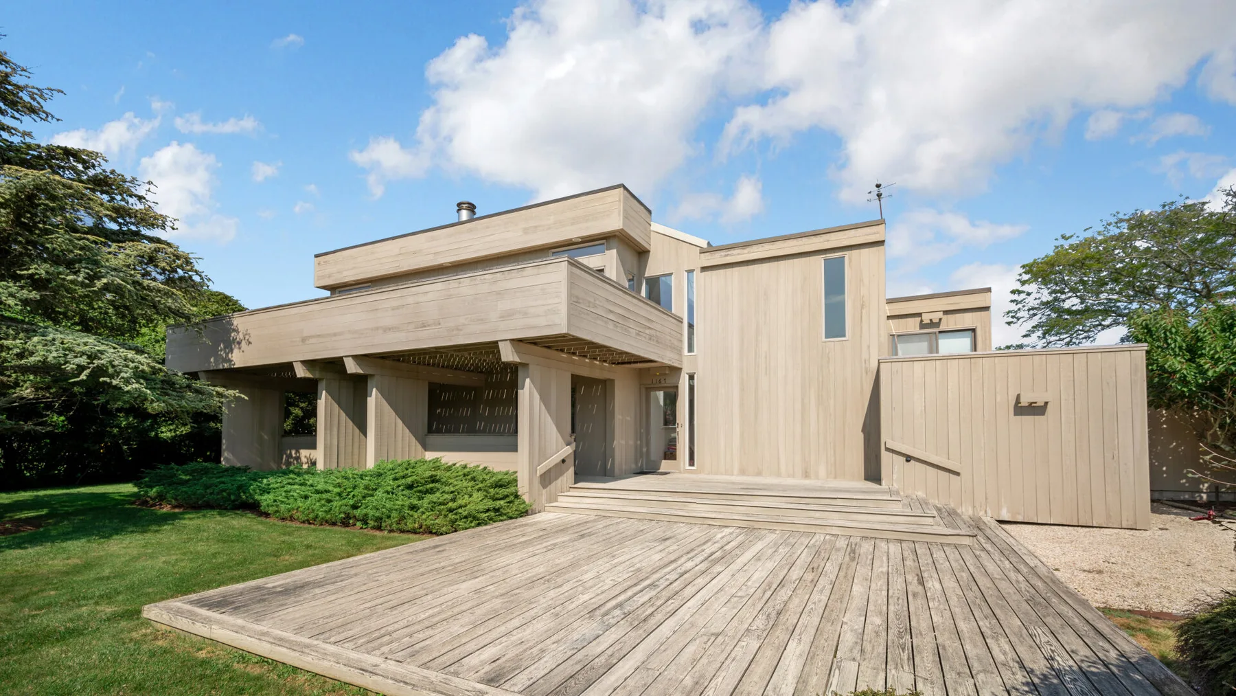 A modern wood-clad house stands behind a broad wooden deck, with trees around it.