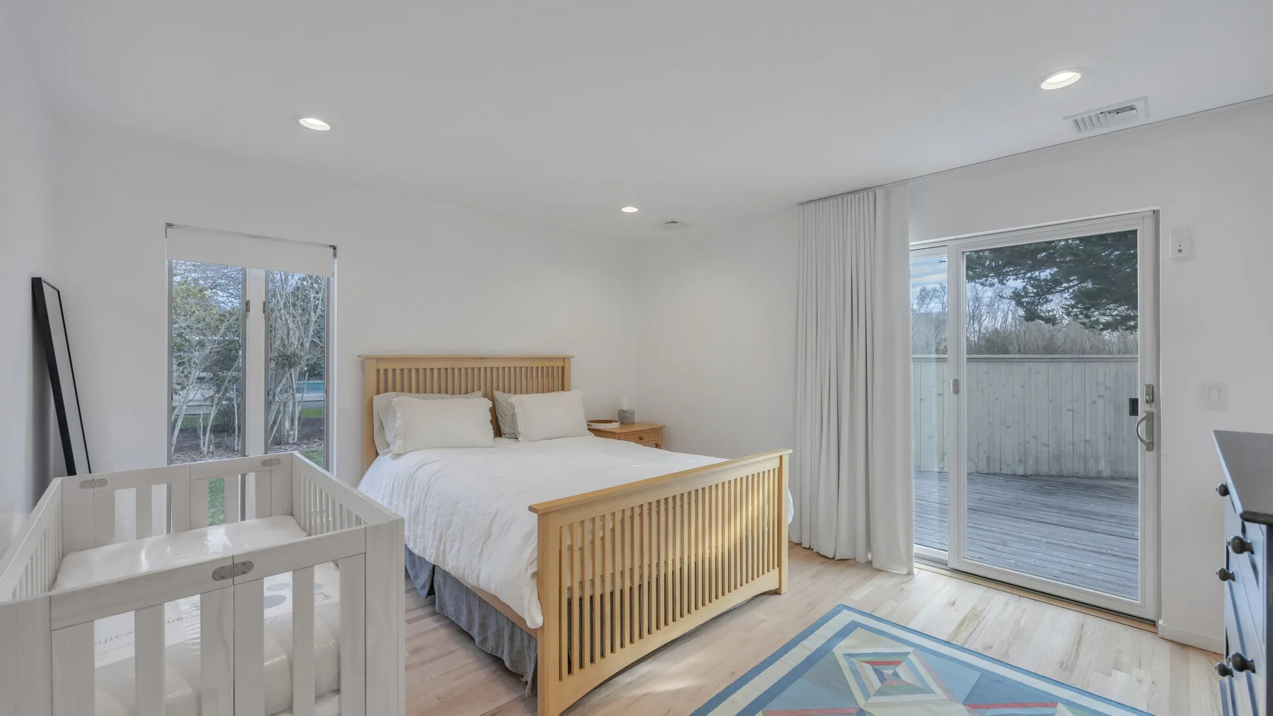 A bright bedroom with a wooden bed, white bedding, and a crib.