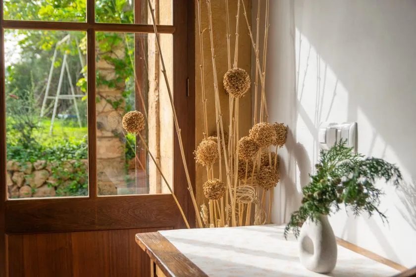 Una mesa de comedor iluminada por el sol, con flores secas junto a una ventana grande.