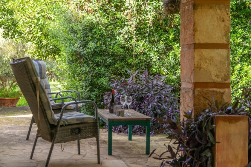 Un patio de jardín con asientos acolchados y una mesa pequeña, rodeado de vegetación.