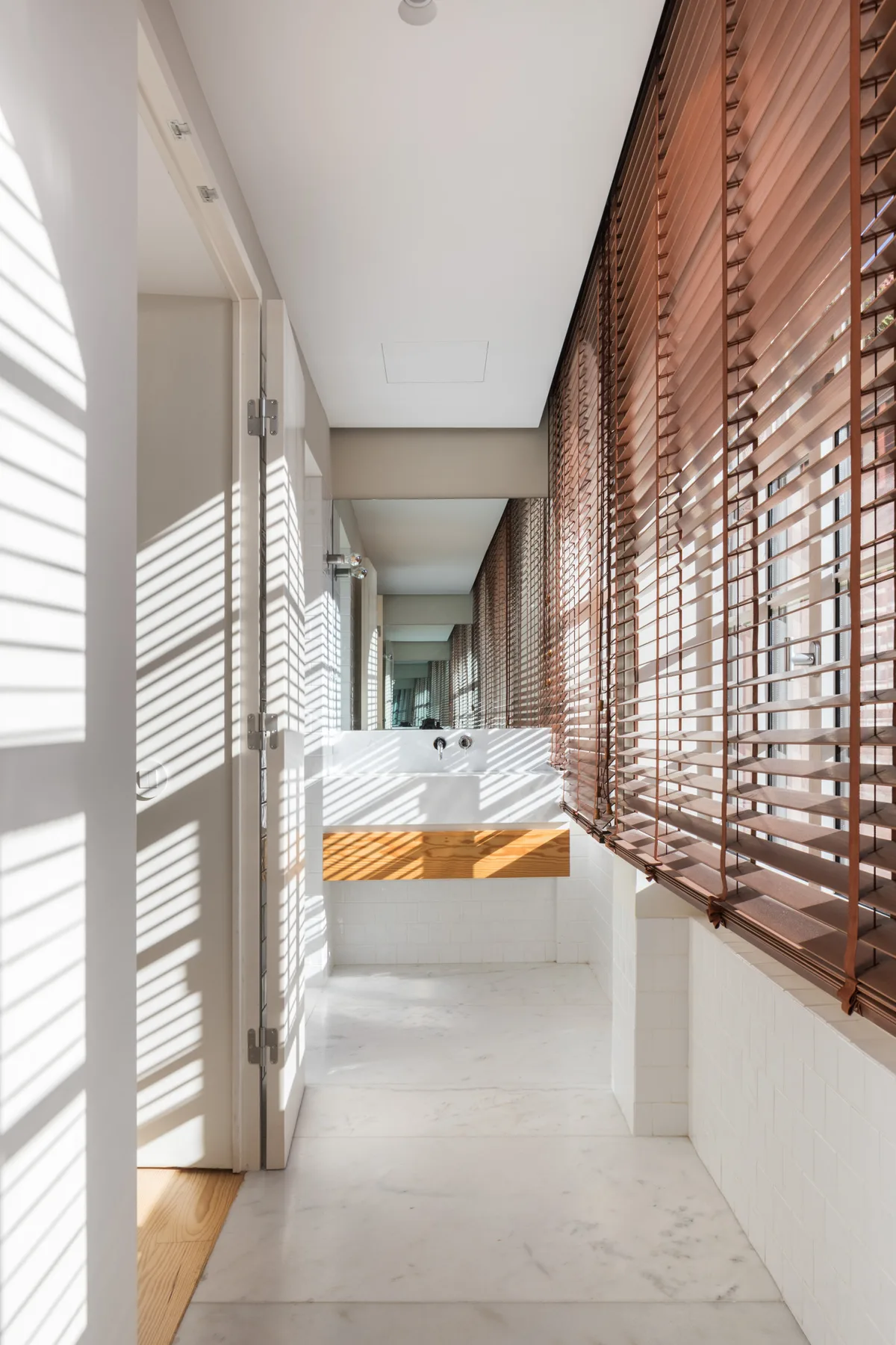 A bright bathroom with a long vanity and sunlight filtering through wooden blinds.