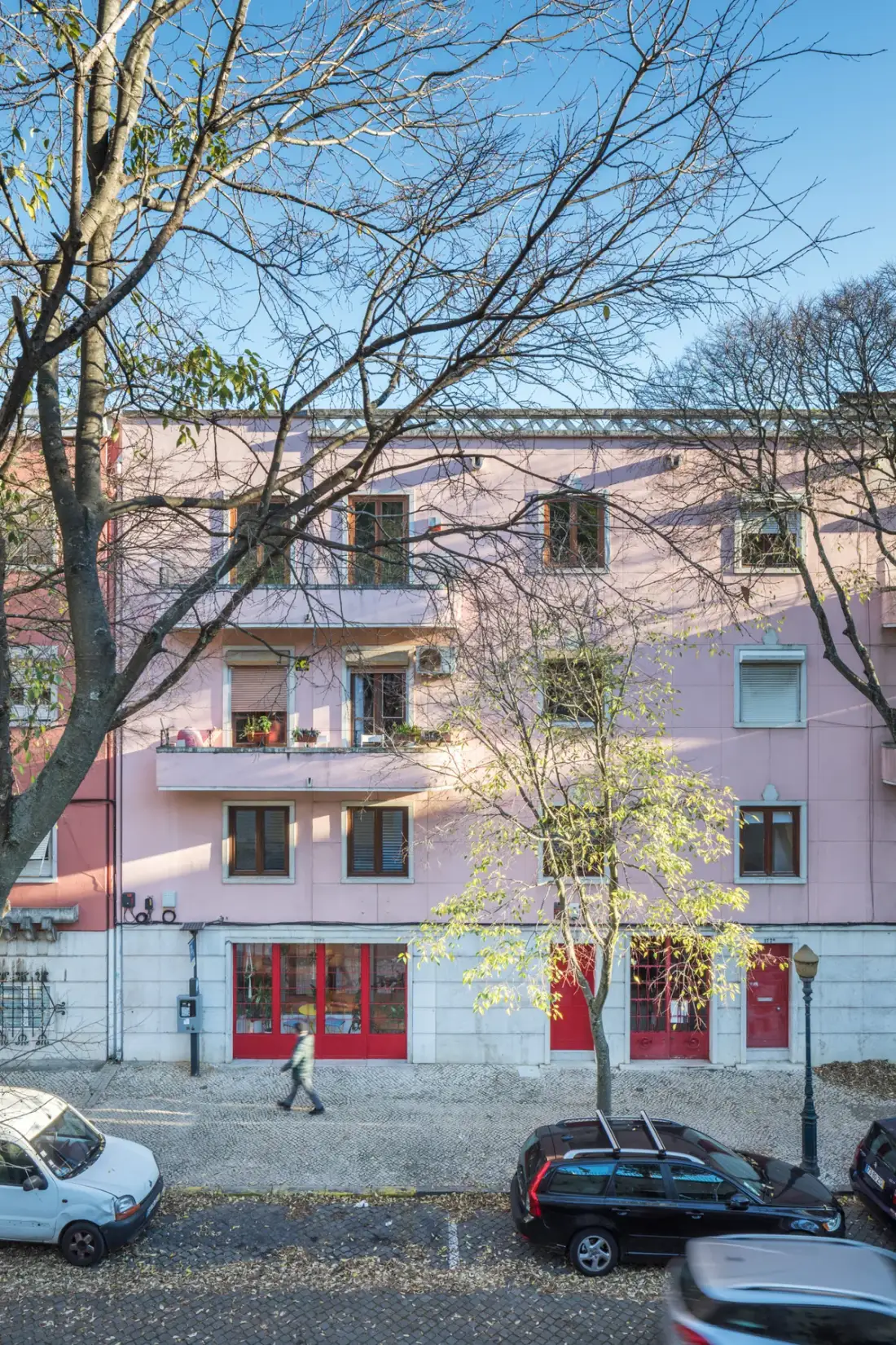 A pink building with balconies and storefronts stands along a tree-lined street.