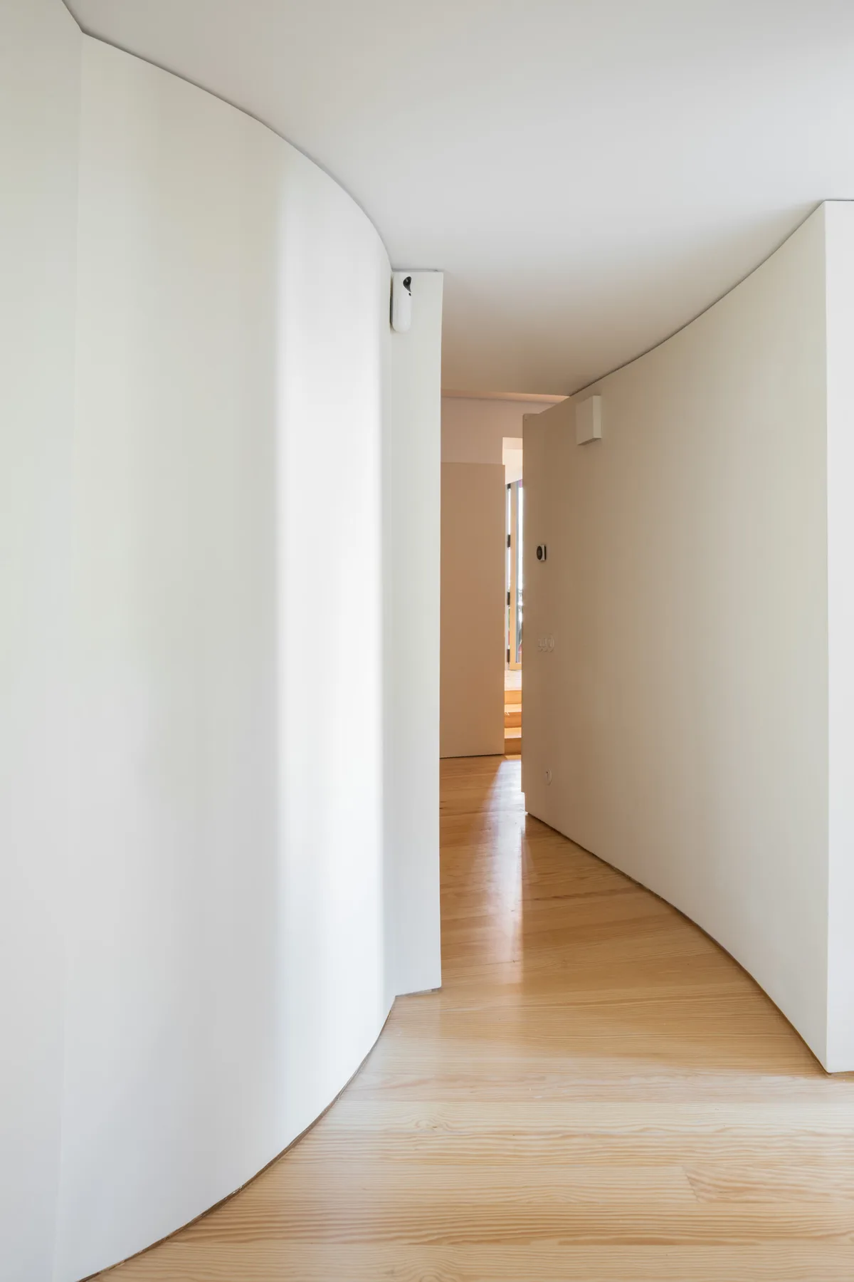 A curved white hallway leads toward a brighter room, with light wood flooring.