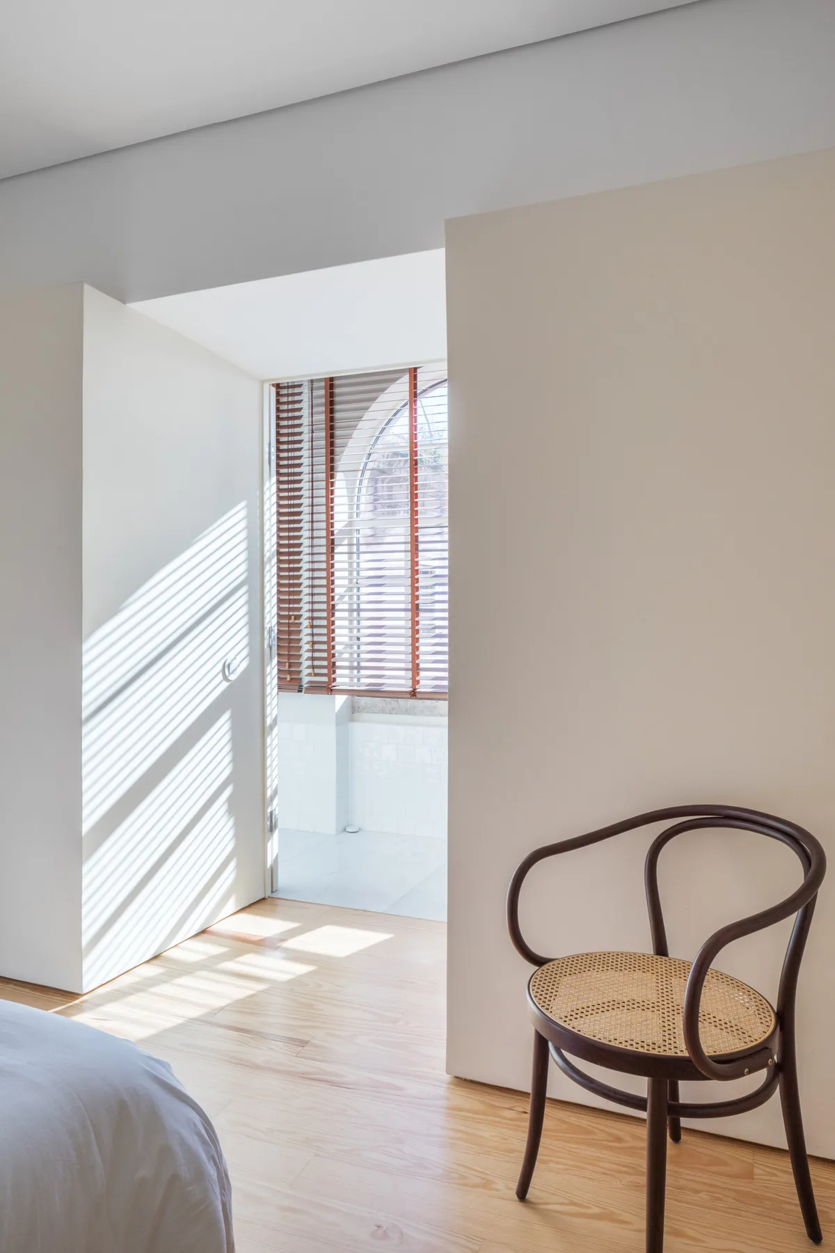 A bright bedroom with a bed, a woven-seat chair, and sunlight through window blinds.