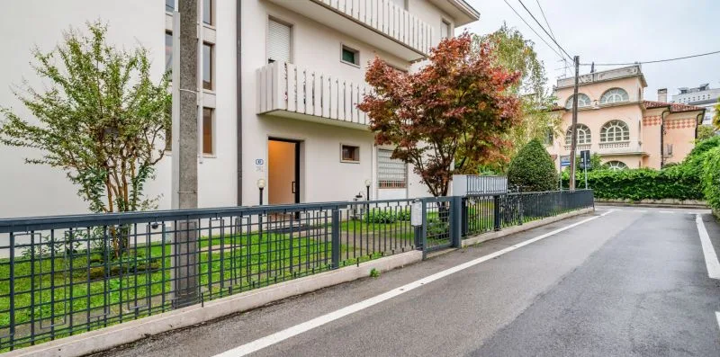 A residential building stands beside a paved street, with trees behind a metal fence.