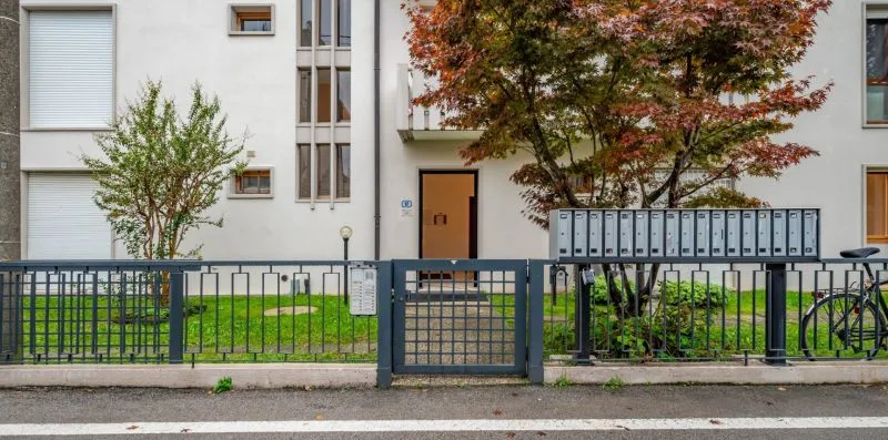 An apartment building entrance behind a metal fence, with trees and a row of mailboxes.