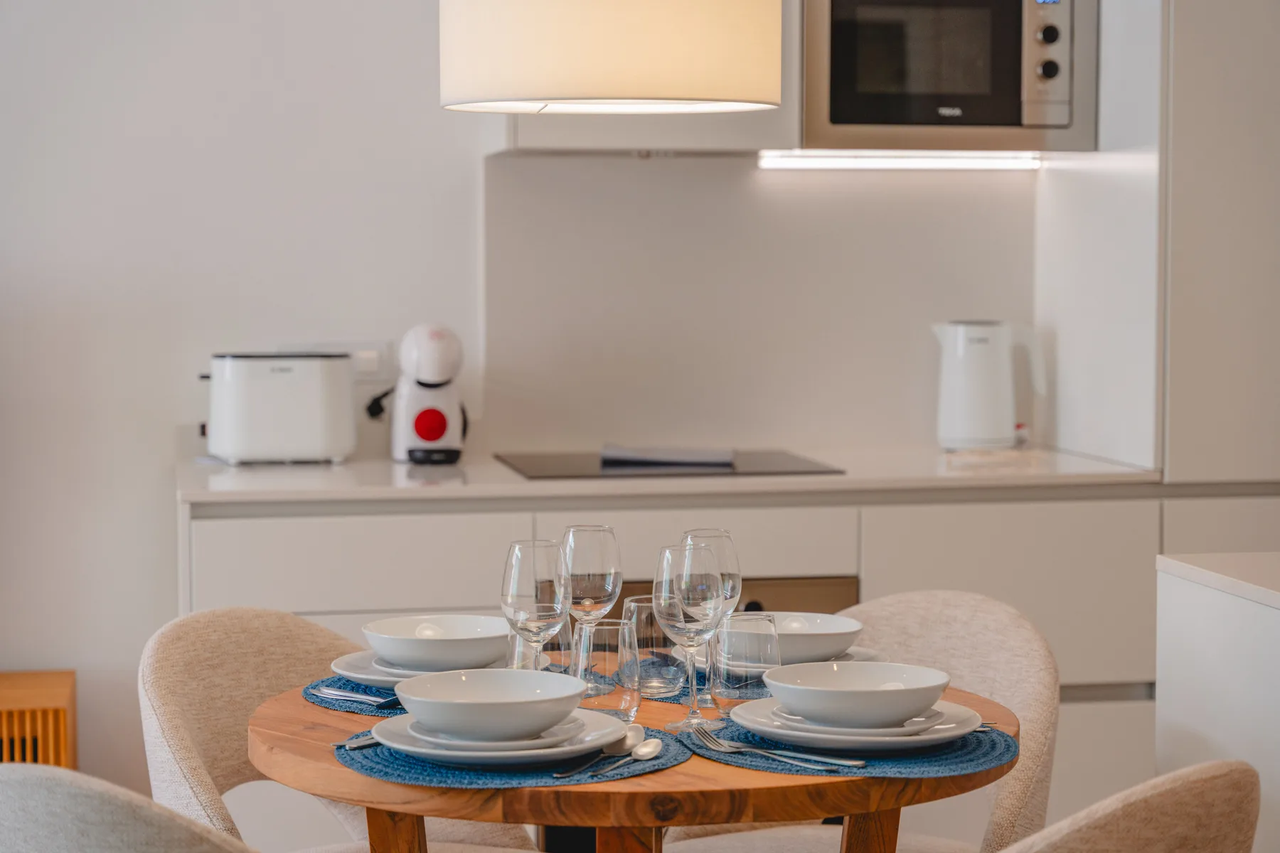 A dining table set with plates and glassware sits in front of a compact kitchen.