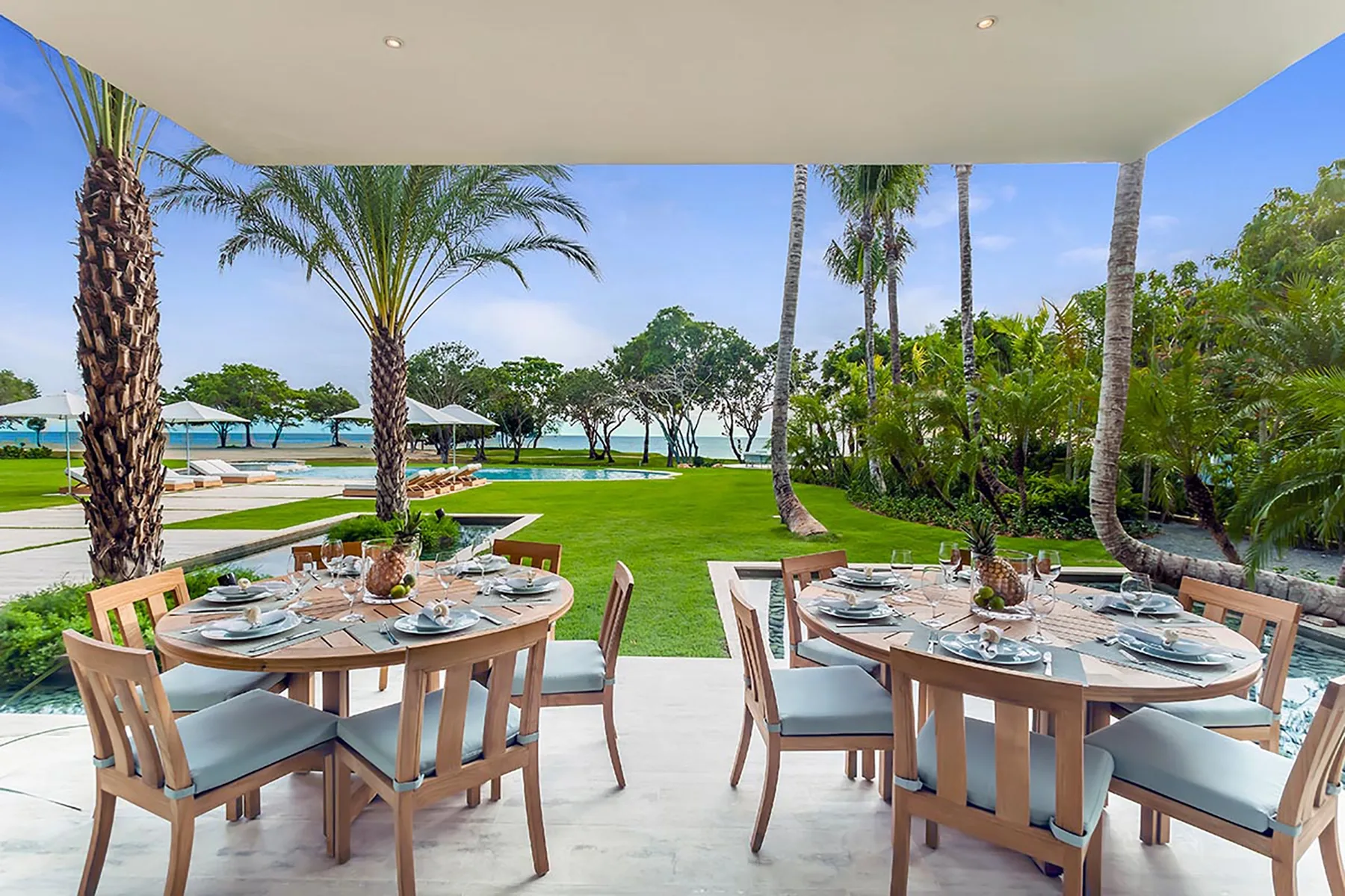 Un comedor al aire libre con mesas preparadas da a un césped, palmeras y el agua.