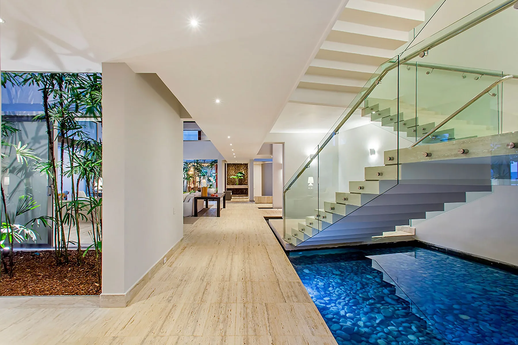 Un pasillo interior luminoso se abre junto a una escalera con barandilla de vidrio y una piscina interior.