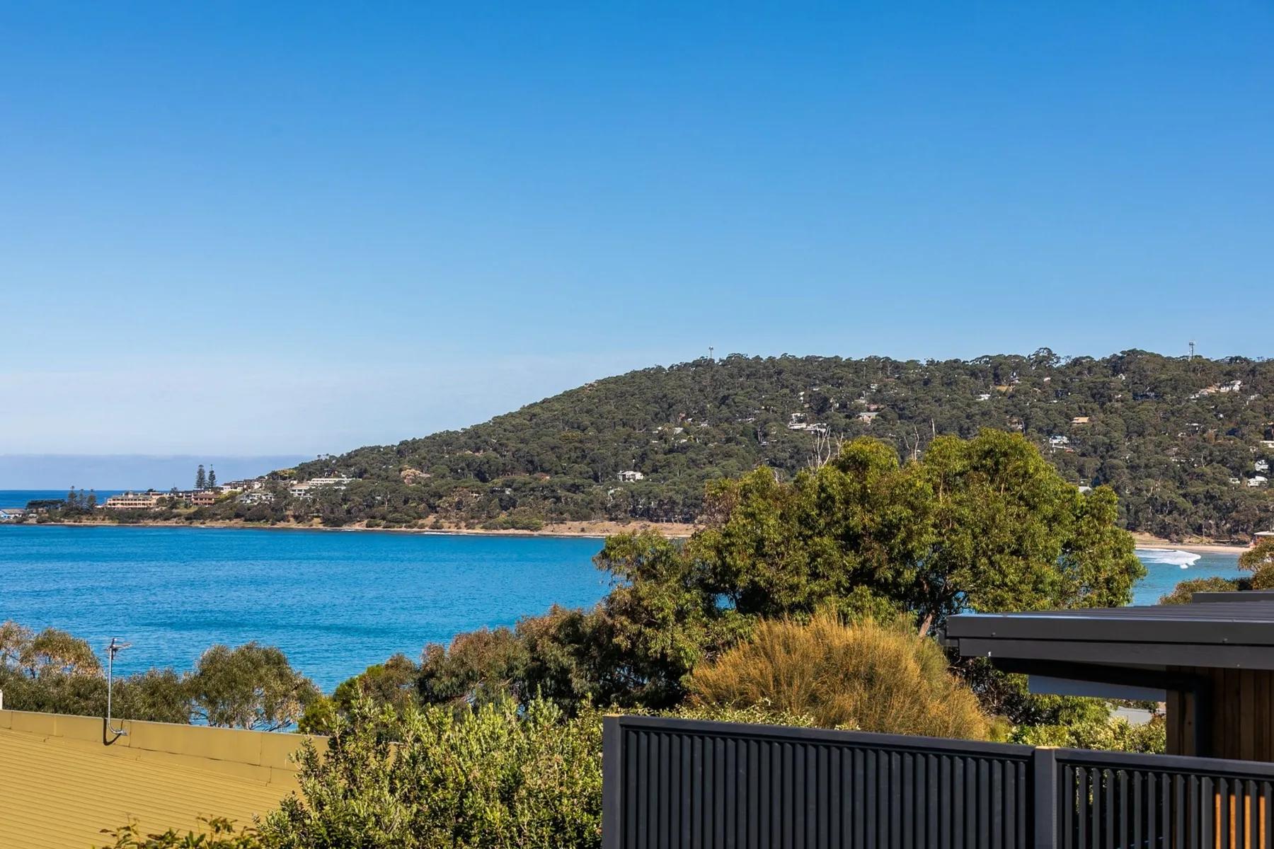 A coastal view shows blue water, a wooded hillside, and a dark balcony railing.