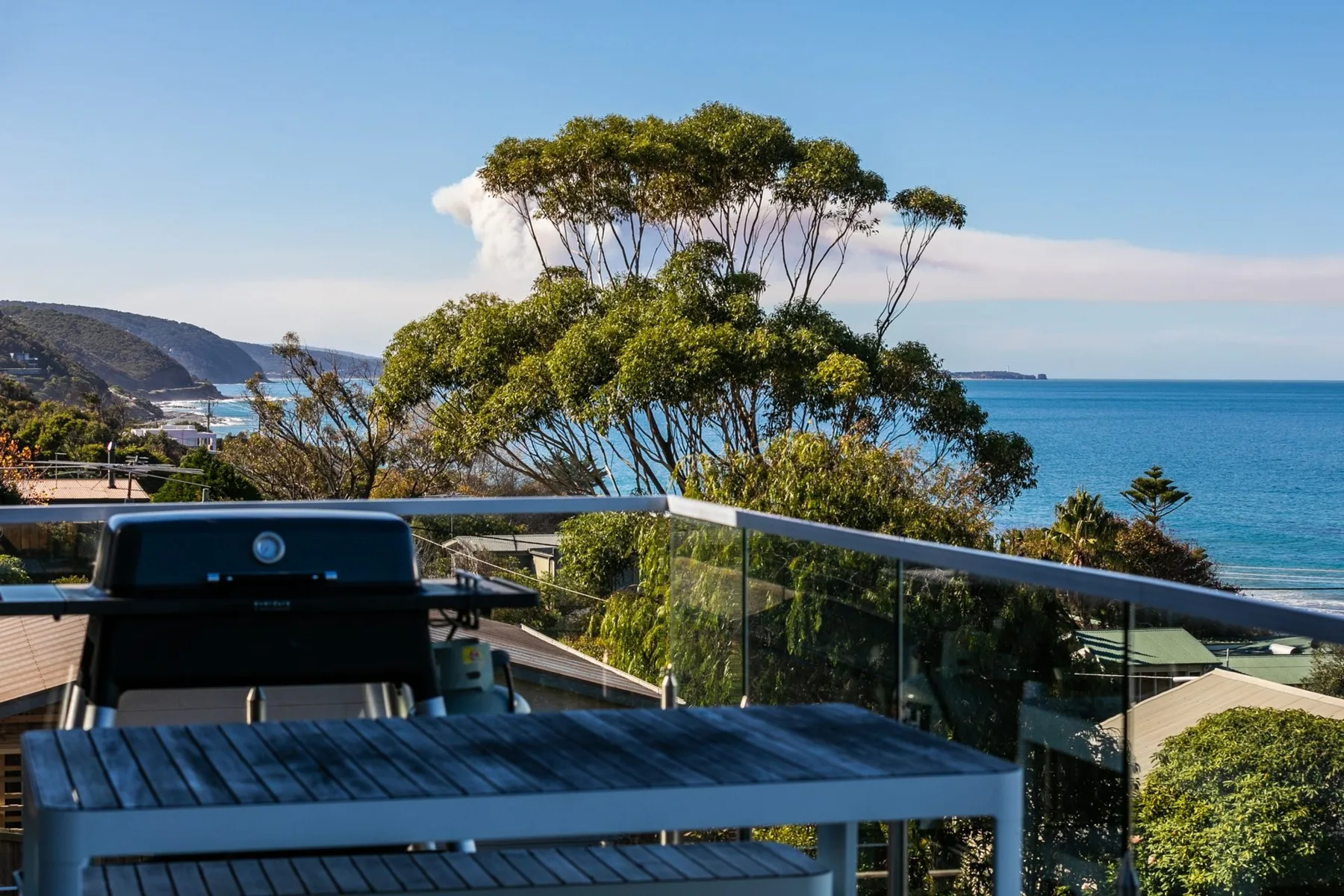 An outdoor dining area overlooks the ocean, with a table and grill beside the glass railing.