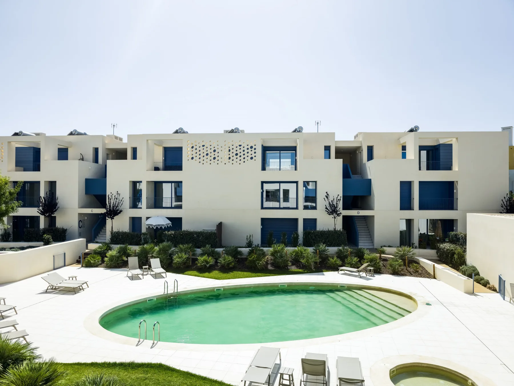 A modern apartment building overlooks a curved swimming pool and surrounding lounge chairs.