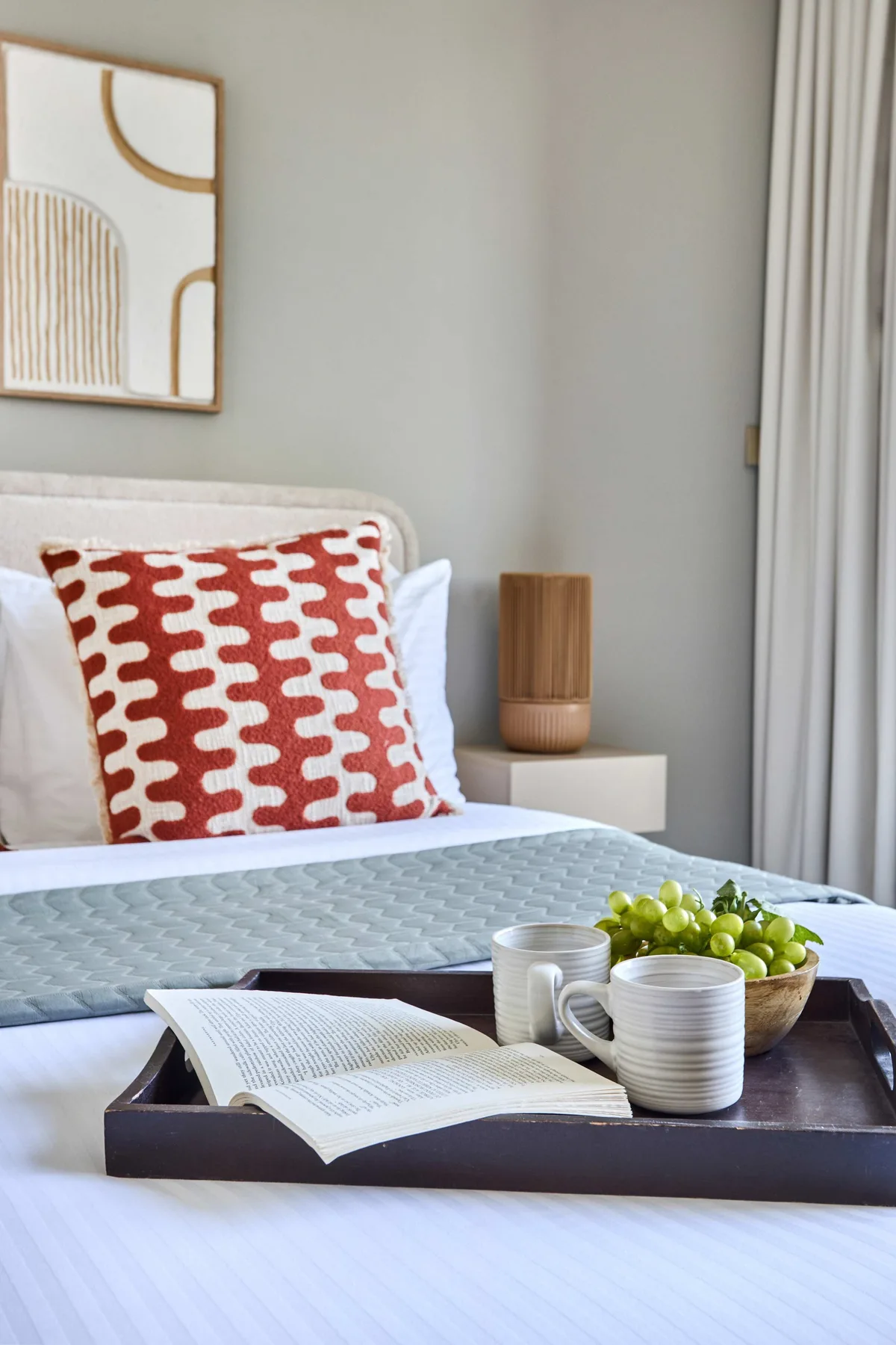 Una cama bien arreglada tiene cojines estampados, un libro abierto y una bandeja con tazas y fruta.