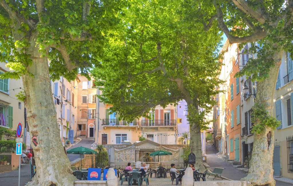 Una cafetería al aire libre, a la sombra de árboles, está entre edificios coloridos.