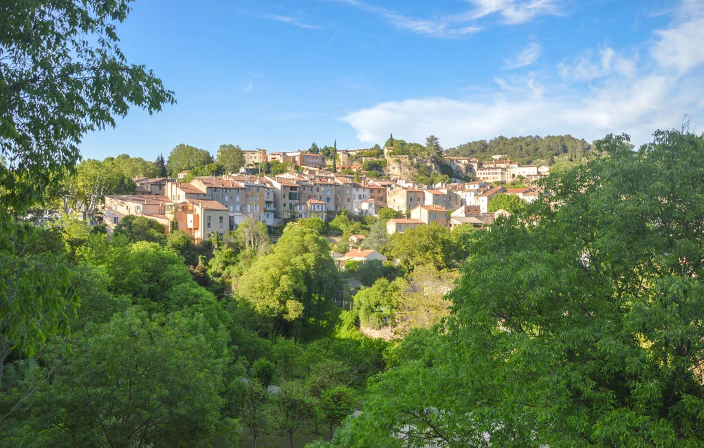 Un pueblo en una ladera, con edificios agrupados, está rodeado de árboles verdes bajo un cielo azul.