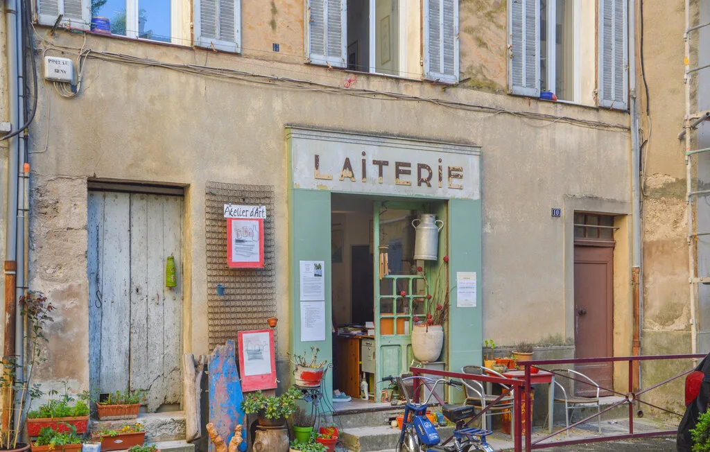 Entrada de una tienda a la calle bajo un letrero que dice «LAITERIE», con plantas y bicicletas afuera.
