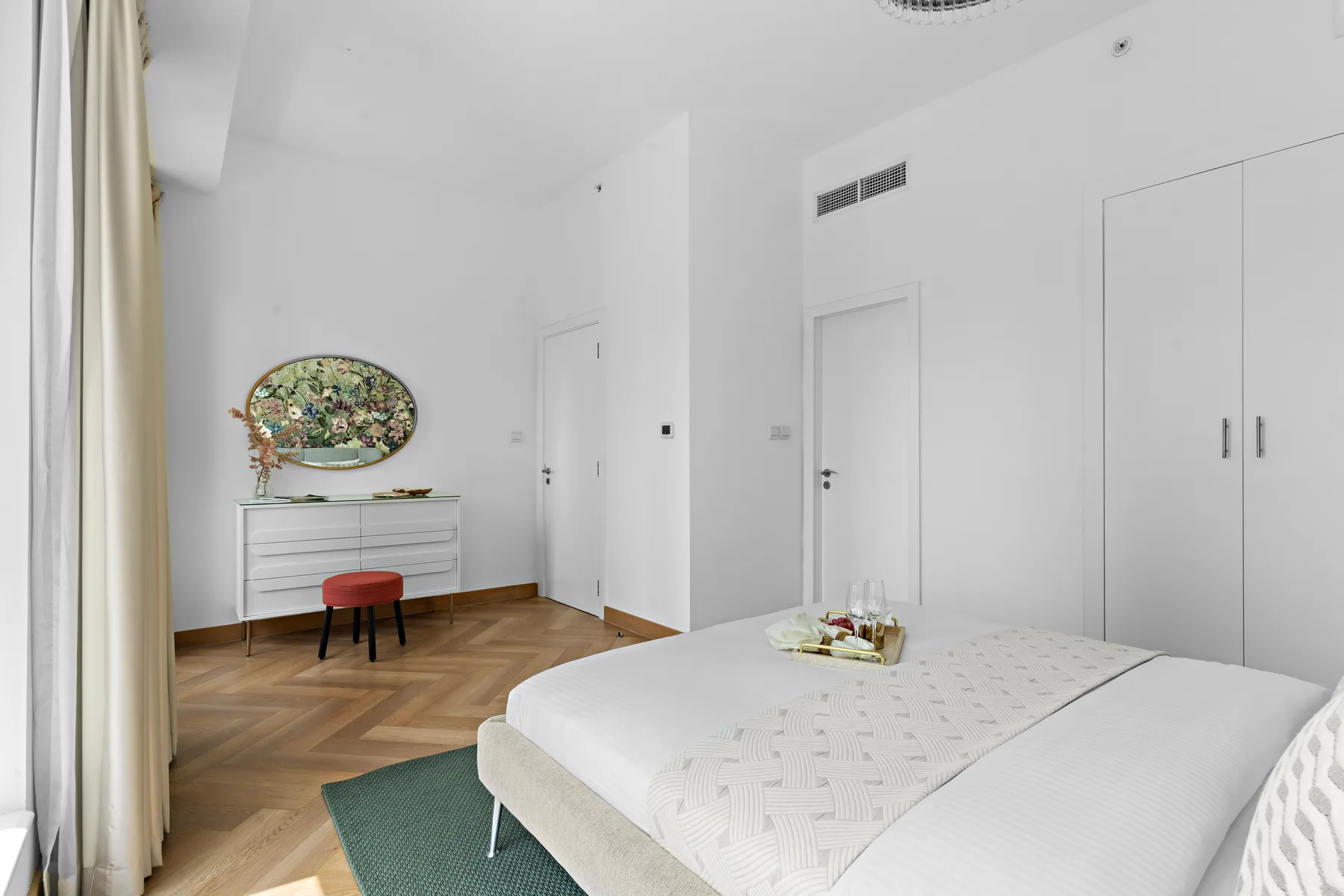 A bright bedroom with a white bed, dresser, and oval mirror.