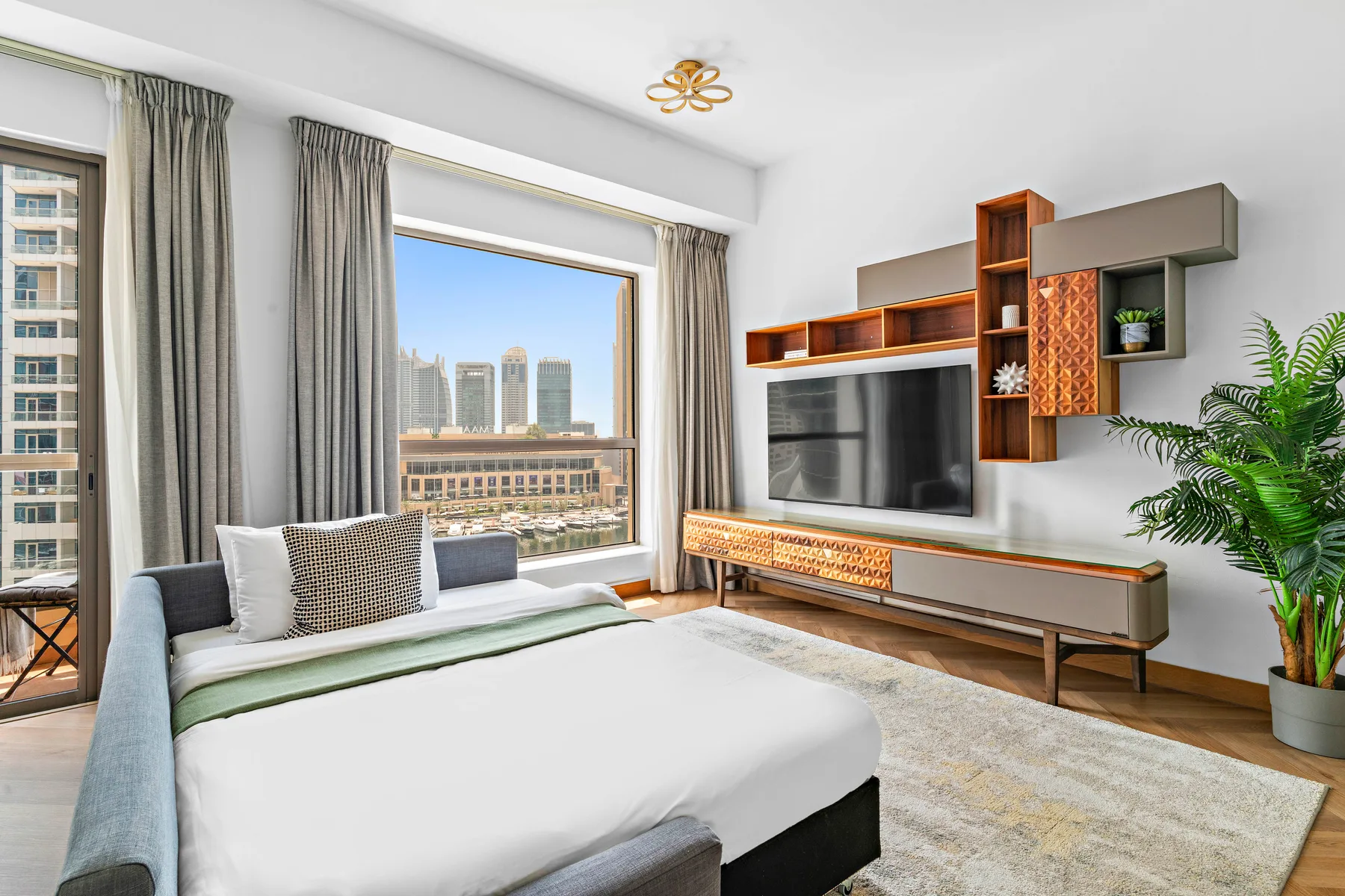 A bright bedroom with a large bed, wall-mounted television, and broad windows.