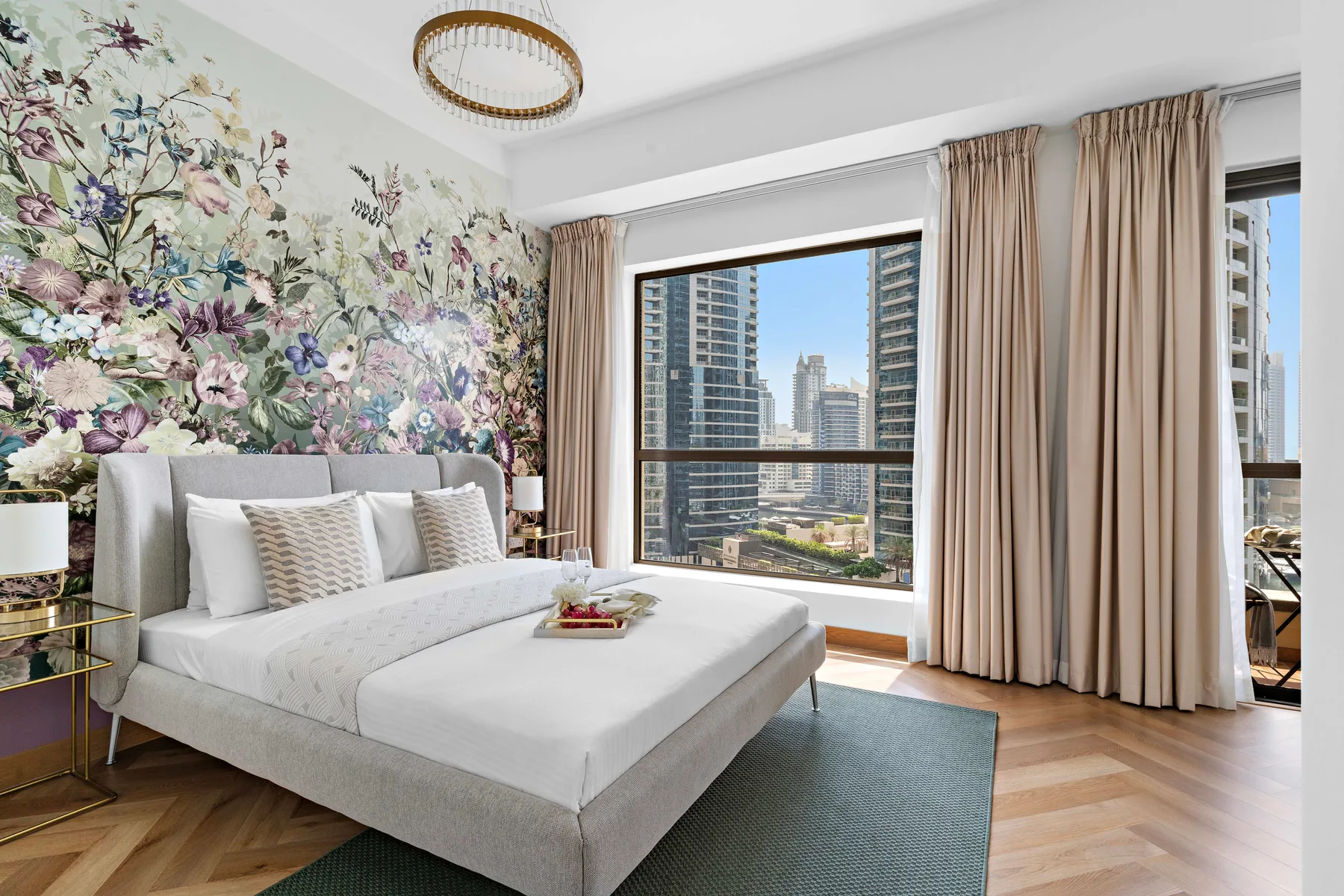 A bedroom with a bed, floral wall covering, and large windows overlooking nearby buildings.