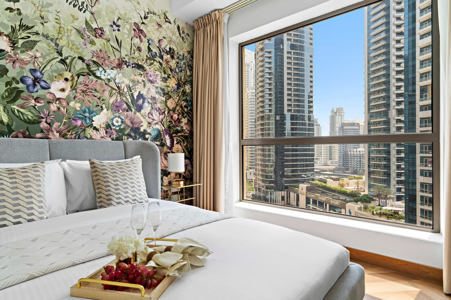 A bedroom with a bed beside a large window overlooking high-rise buildings.
