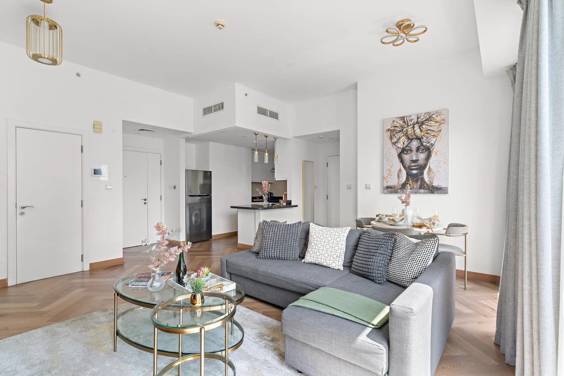 A bright living room with gray seating, glass coffee tables, and an open kitchen.