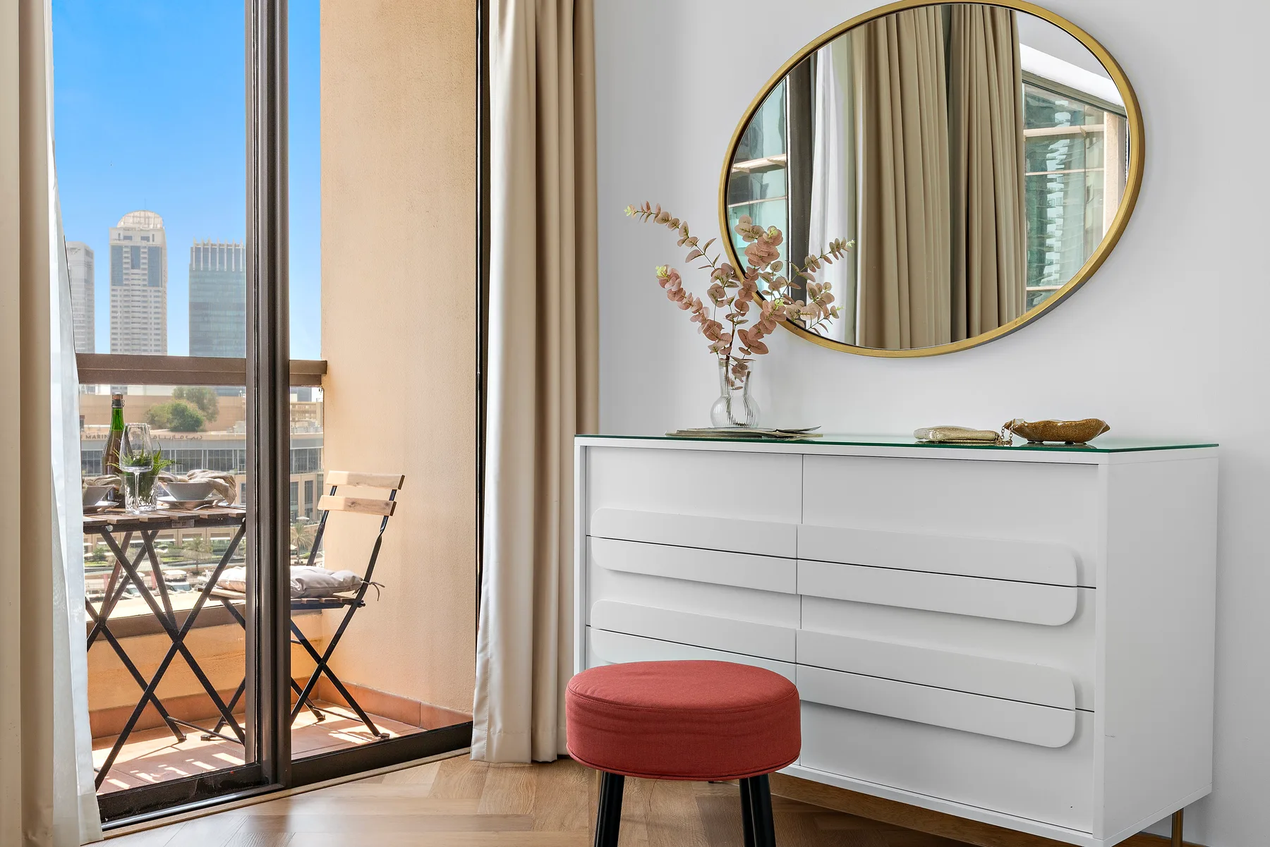 A bright bedroom seating area with a white dresser and oval mirror beside a balcony.