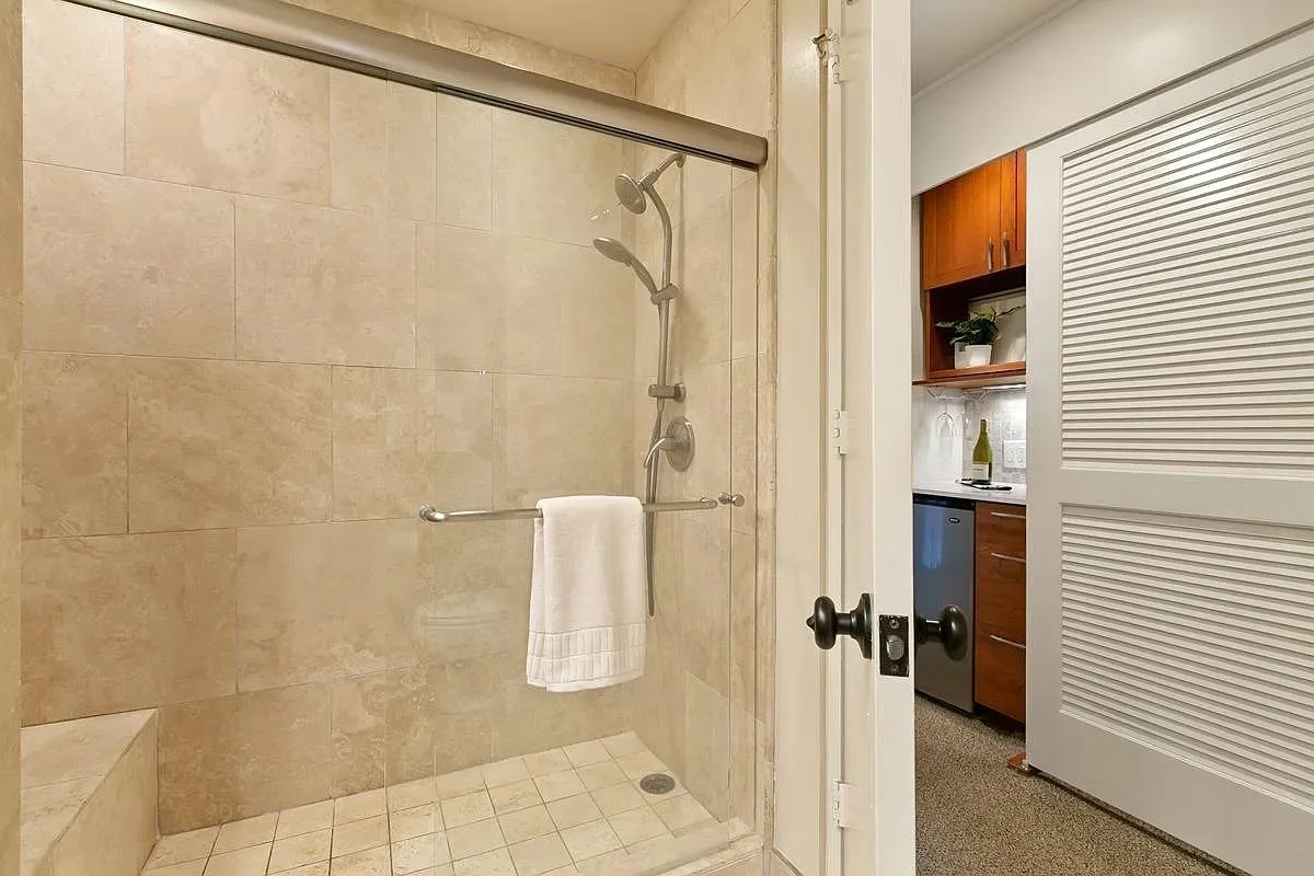 Se ve una ducha de baño con azulejos junto a una puerta abierta a una pequeña cocina.