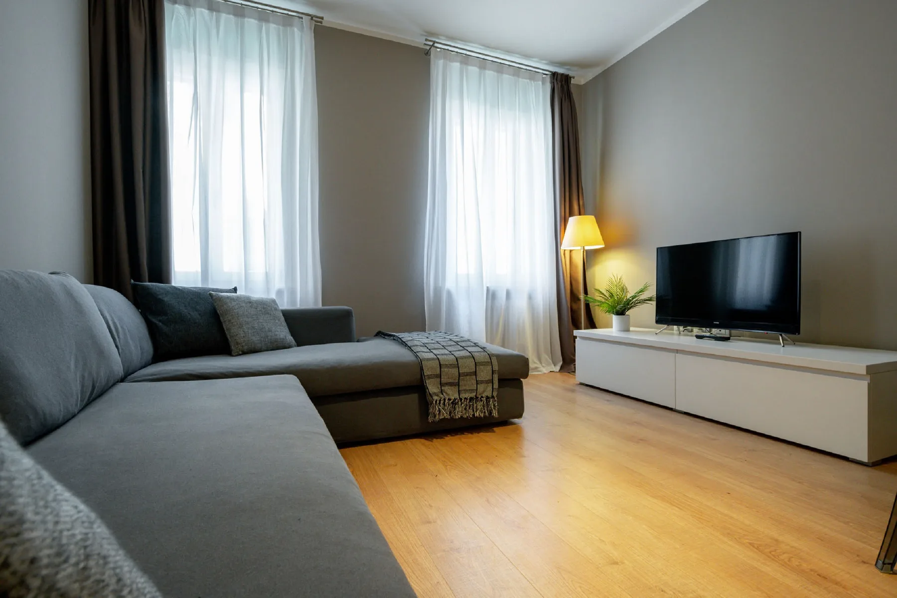 Una sala de estar con asientos grises, un televisor y ventanas altas con cortinas.