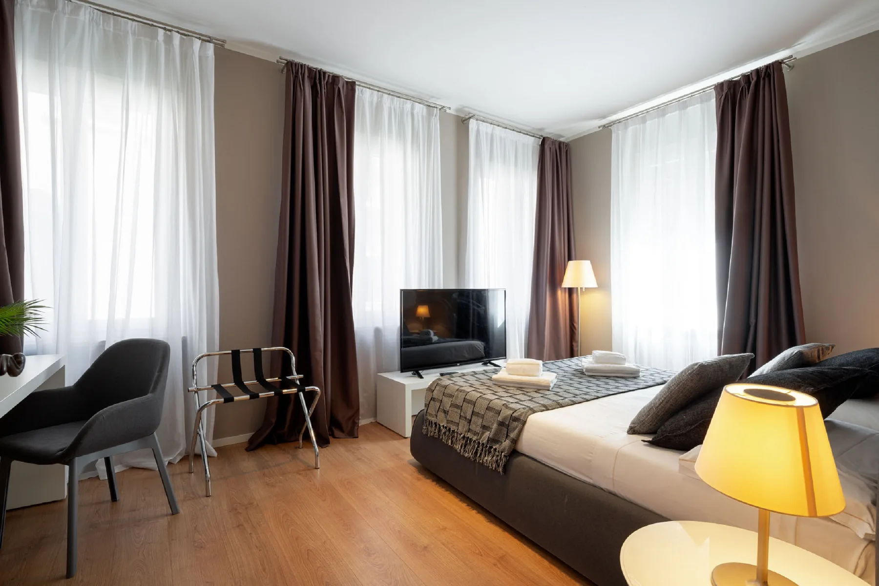 Un dormitorio con cama, televisor y ventanas altas con cortinas.
