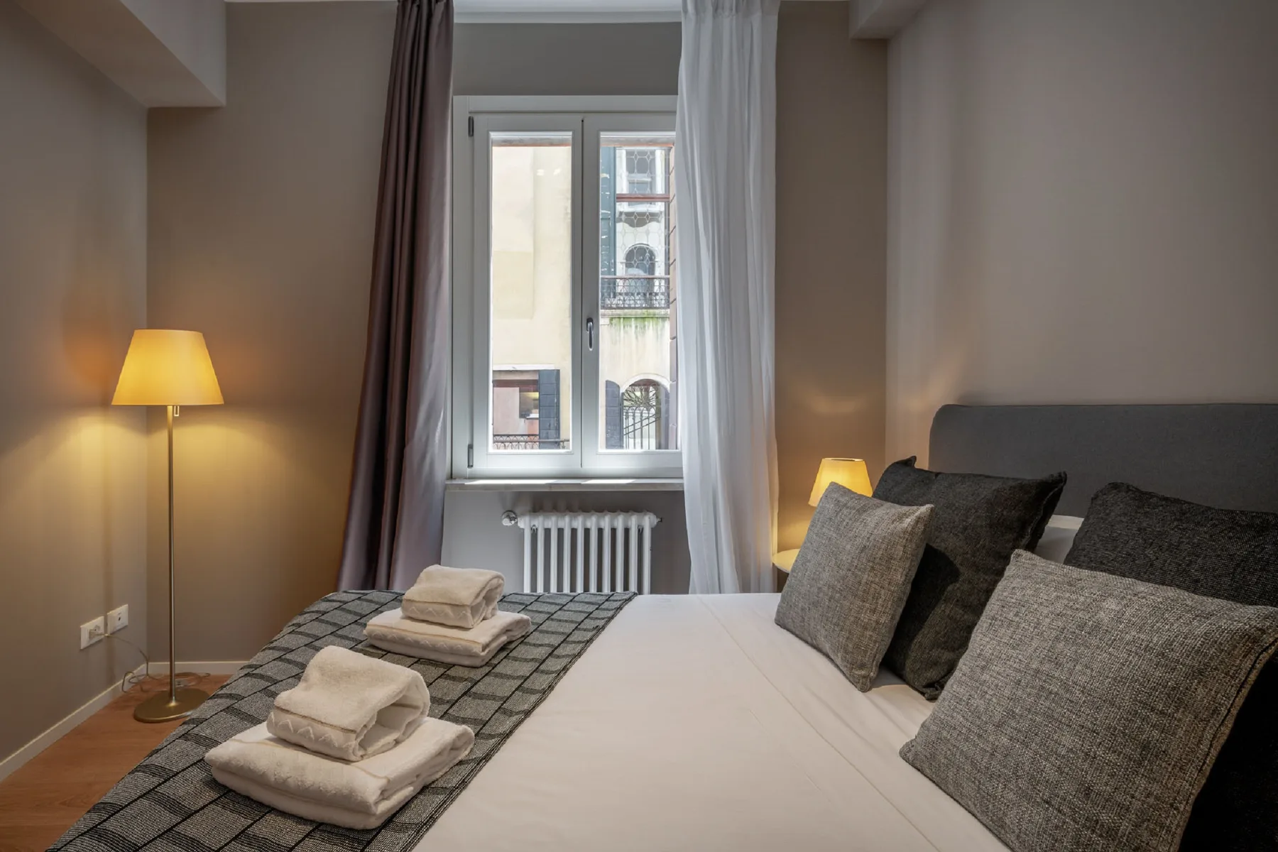 Un dormitorio con una cama, lámparas y una ventana con edificios cercanos a la vista.