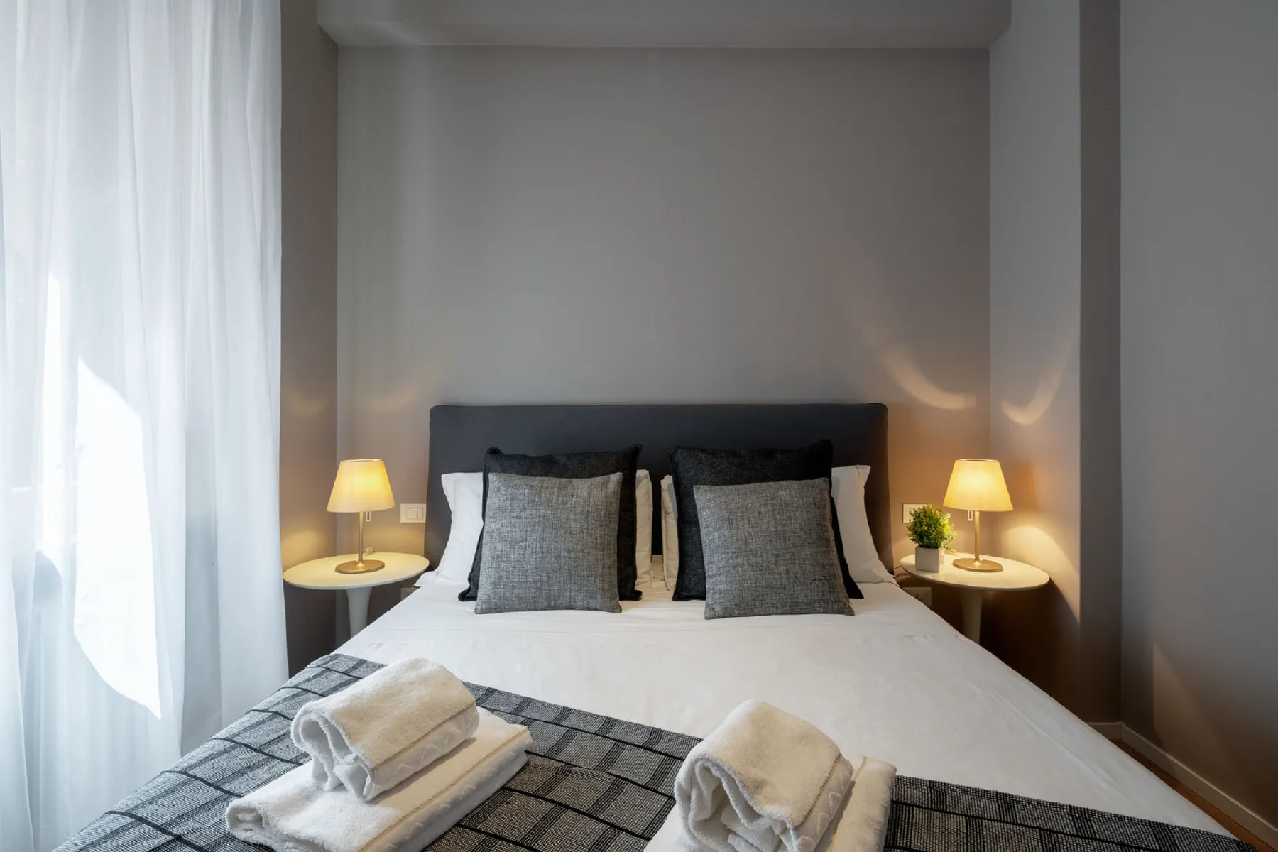 Un dormitorio con una cama bien tendida, lámparas de noche y cortinas translúcidas.