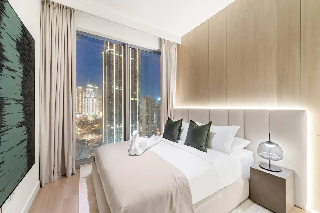 A bedroom with a large bed beside floor-to-ceiling windows overlooking a city at night.
