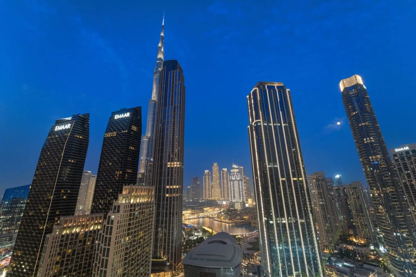 A city skyline at dusk features tall illuminated skyscrapers beneath a blue sky.
